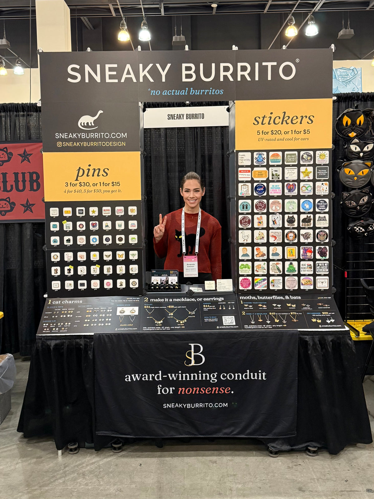 A booth at an indoor event with a person standing behind a display table. The backdrop reads 'SNEAKY BURRITO' with product sections for pins, stickers, and burrito coins, and the table has jewelry and small items displayed.