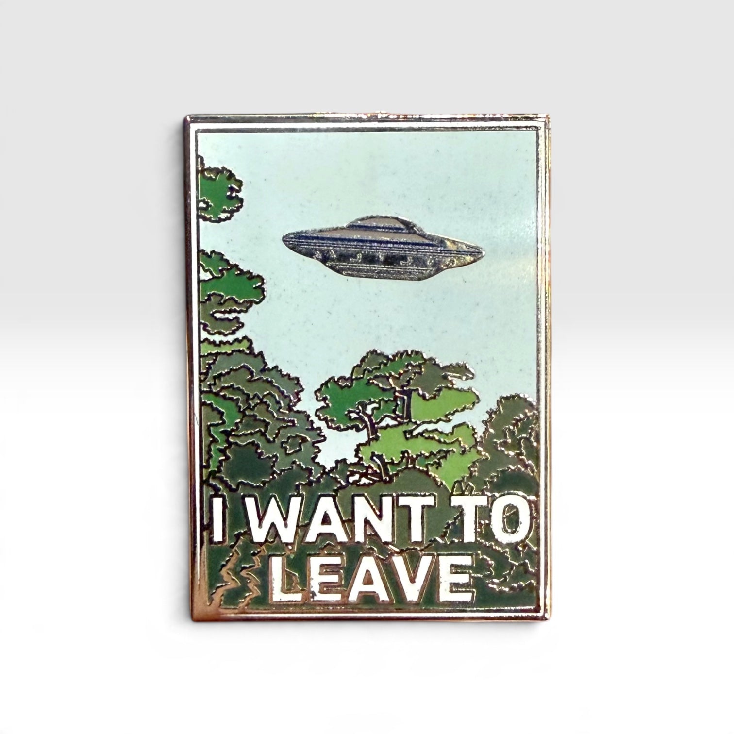 A rectangular enamel pin with a vintage-style border, featuring a light blue sky, green trees, a silver-gray UFO, and the text 'I WANT TO LEAVE' in white. The pin is photographed against a clean, light gray gradient background.