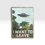 A rectangular enamel pin with a vintage-style border, featuring a light blue sky, green trees, a silver-gray UFO, and the text 'I WANT TO LEAVE' in white. The pin is photographed against a clean, light gray gradient background.