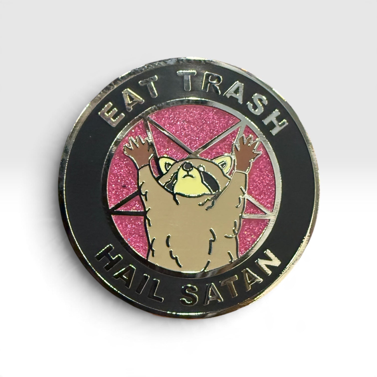 A circular enamel pin featuring a raccoon with raised paws on a pink glitter background, surrounded by a black ring with the text 'EAT TRASH' at the top and 'HAIL SATAN' at the bottom.