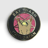 A circular enamel pin featuring a raccoon with raised paws on a pink glitter background, surrounded by a black ring with the text 'EAT TRASH' at the top and 'HAIL SATAN' at the bottom.