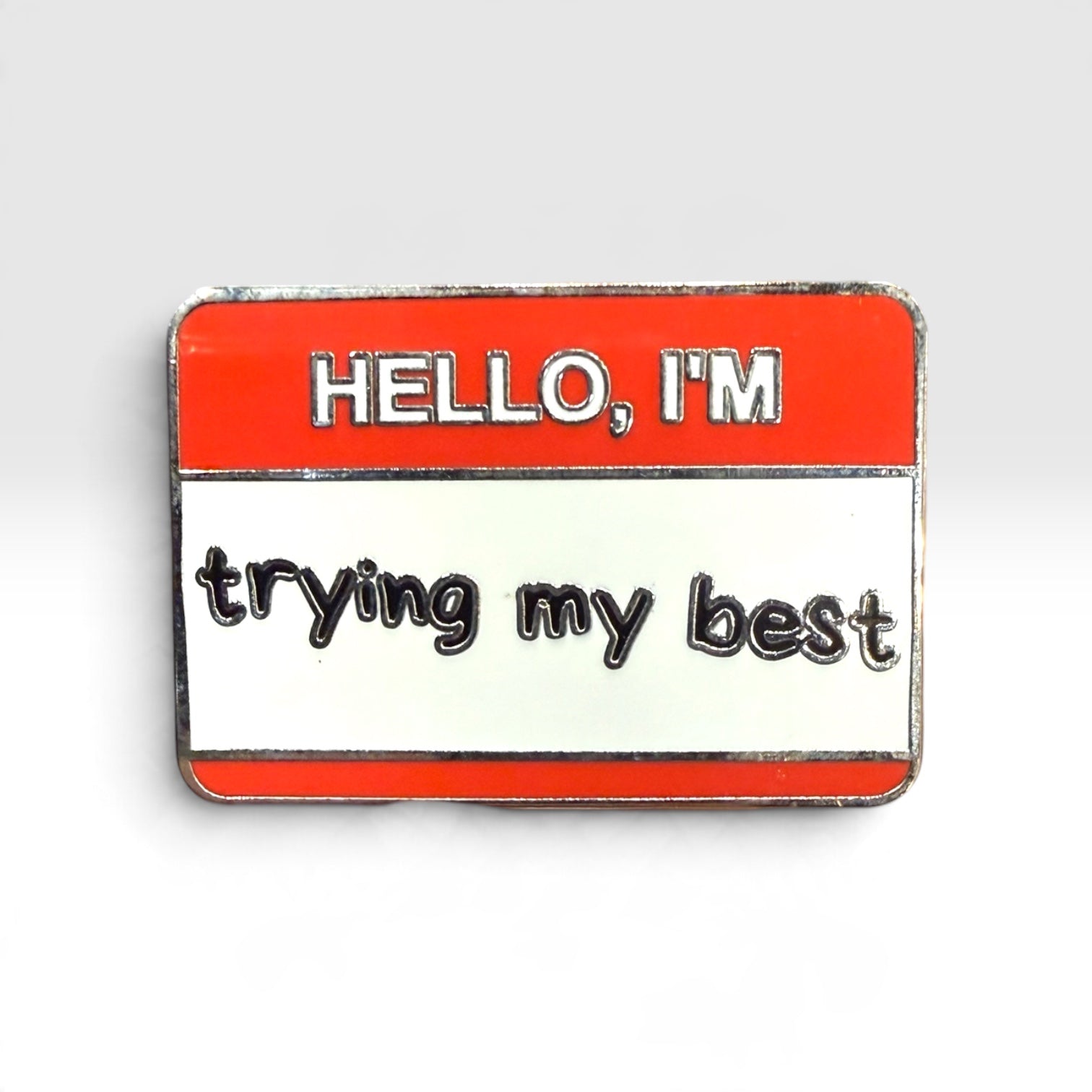 A rectangular enamel pin designed like a name tag, with a red top section reading 'HELLO, I'M' and a white bottom section reading 'trying my best' in black script, outlined in silver metal.