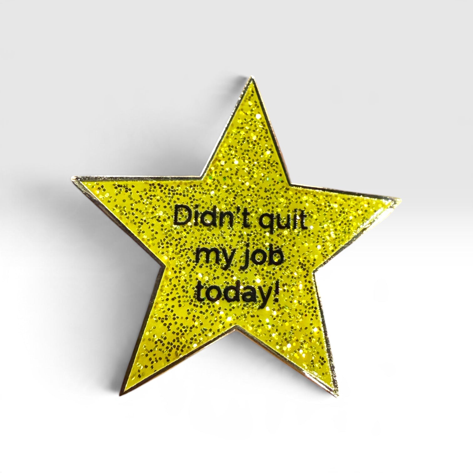 A glittery yellow star-shaped enamel pin with black text reading 'Didn't quit my job today!' on a light gray background.