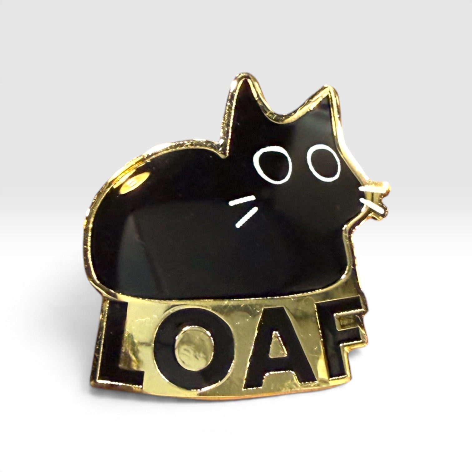 A black cat-shaped enamel pin with gold trim and the word 'LOAF' in black on a gold banner below, shown on a light gray background.