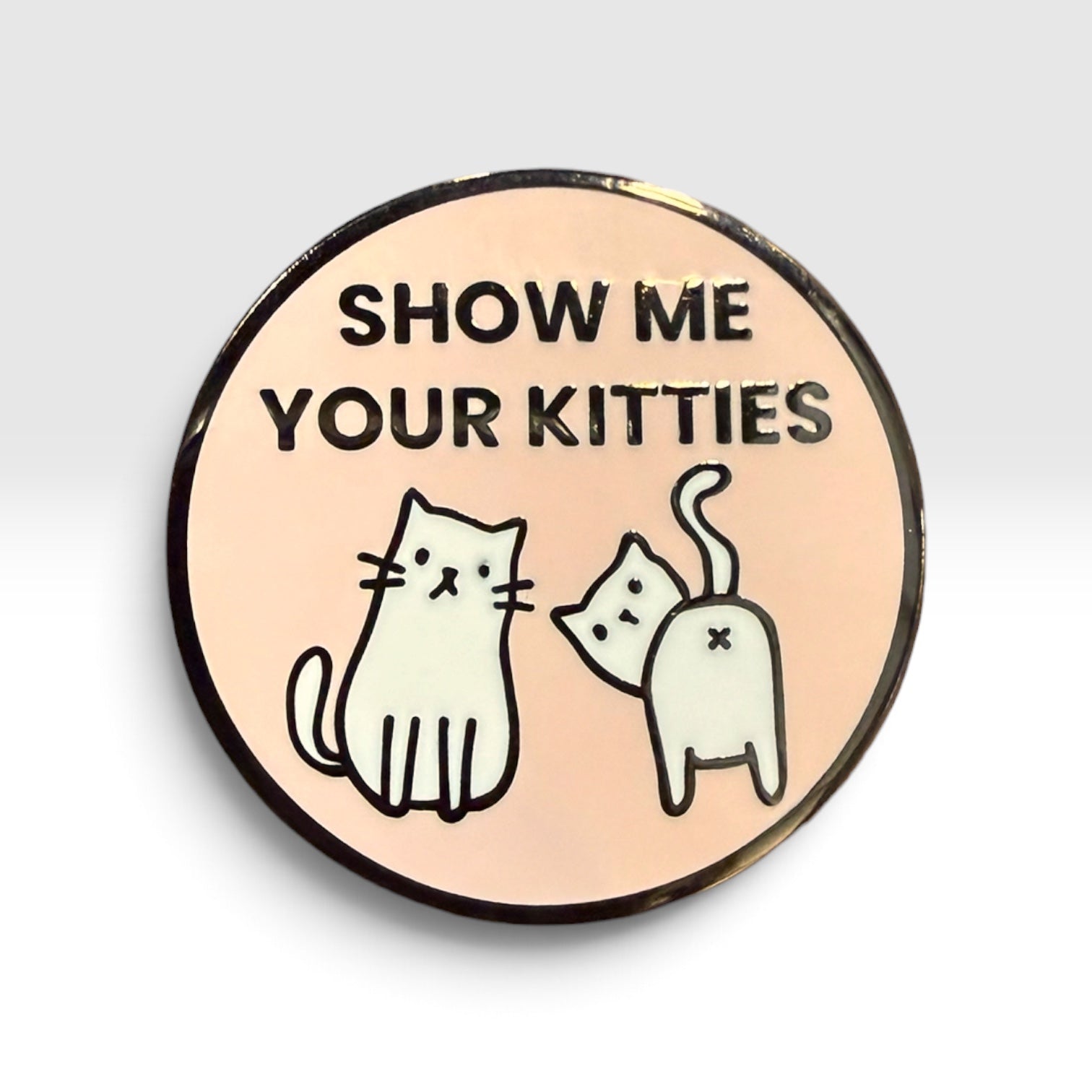 A round enamel pin with a peach-pink background, featuring the black text 'SHOW ME YOUR KITTIES' above two simple black-outlined white cat illustrations. The pin has a dark metallic rim and is photographed on a light gray background.