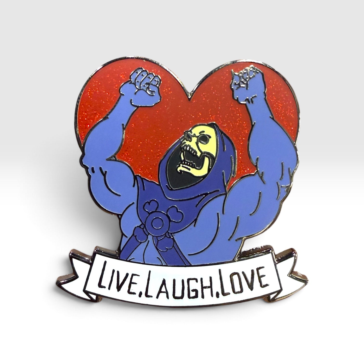 An enamel pin featuring a cartoon skeleton with raised fists in front of a red glitter heart, with a white banner below reading 'LIVE.LAUGH.LOVE.'