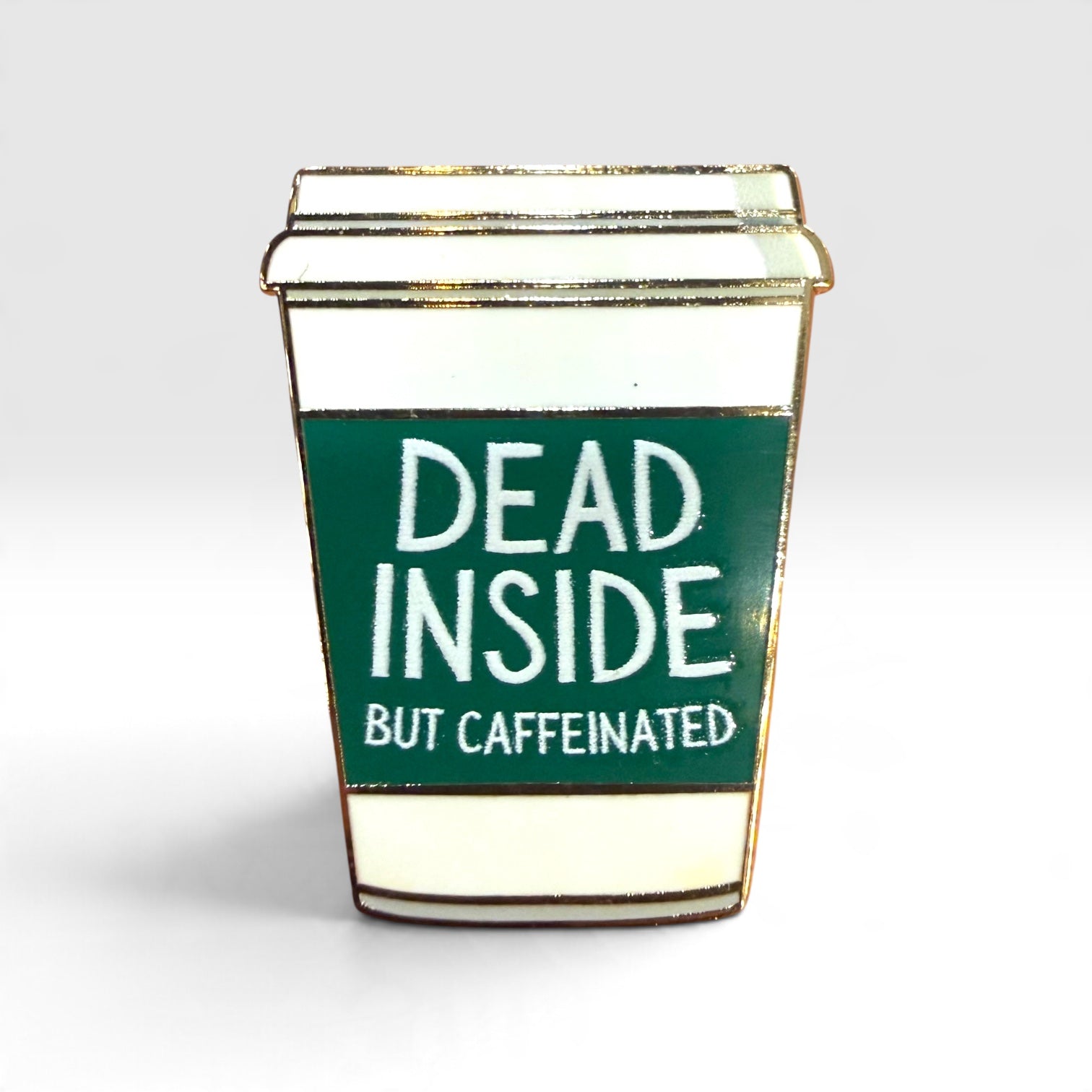 A novelty enamel pin shaped like a takeaway coffee cup with a dark green label that reads 'DEAD INSIDE BUT CAFFEINATED' in white text, outlined in gold metal on a light gray background.