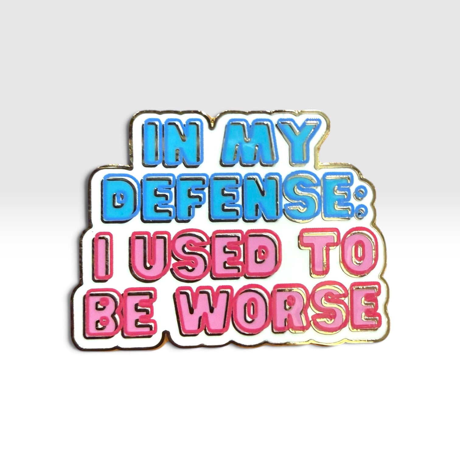 A die-cut enamel pin with the text 'IN MY DEFENSE: I USED TO BE WORSE' in blue and pink bubble letters with a white outline and gold metal border, shown on a light gray gradient background.
