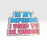 A die-cut enamel pin with the text 'IN MY DEFENSE: I USED TO BE WORSE' in blue and pink bubble letters with a white outline and gold metal border, shown on a light gray gradient background.