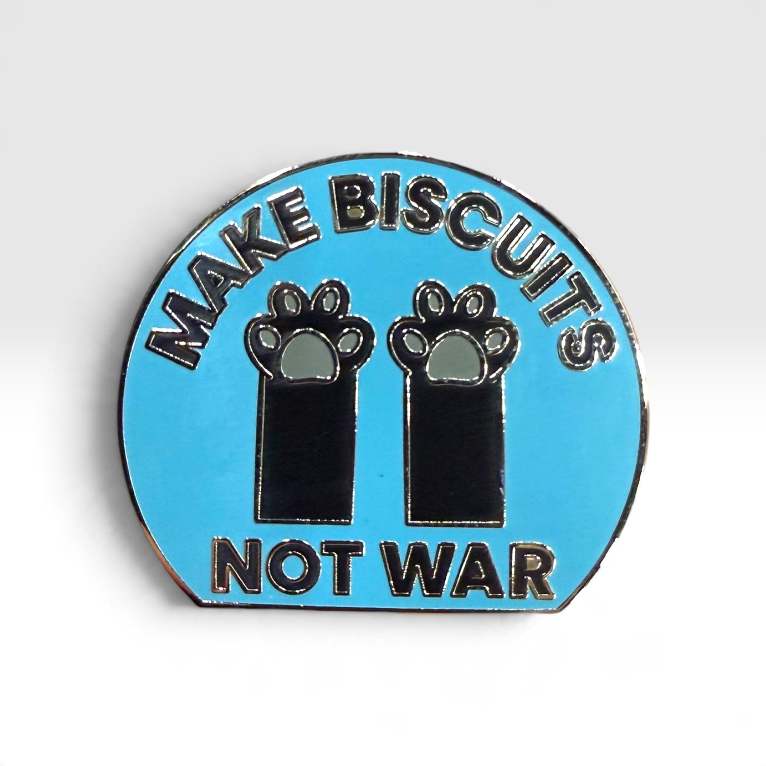 A circular enamel pin with a light blue background, featuring the text 'MAKE BISCUITS NOT WAR' in black lettering with silver outlines, and two black paw prints in the center.