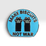A circular enamel pin with a light blue background, featuring the text 'MAKE BISCUITS NOT WAR' in black lettering with silver outlines, and two black paw prints in the center.
