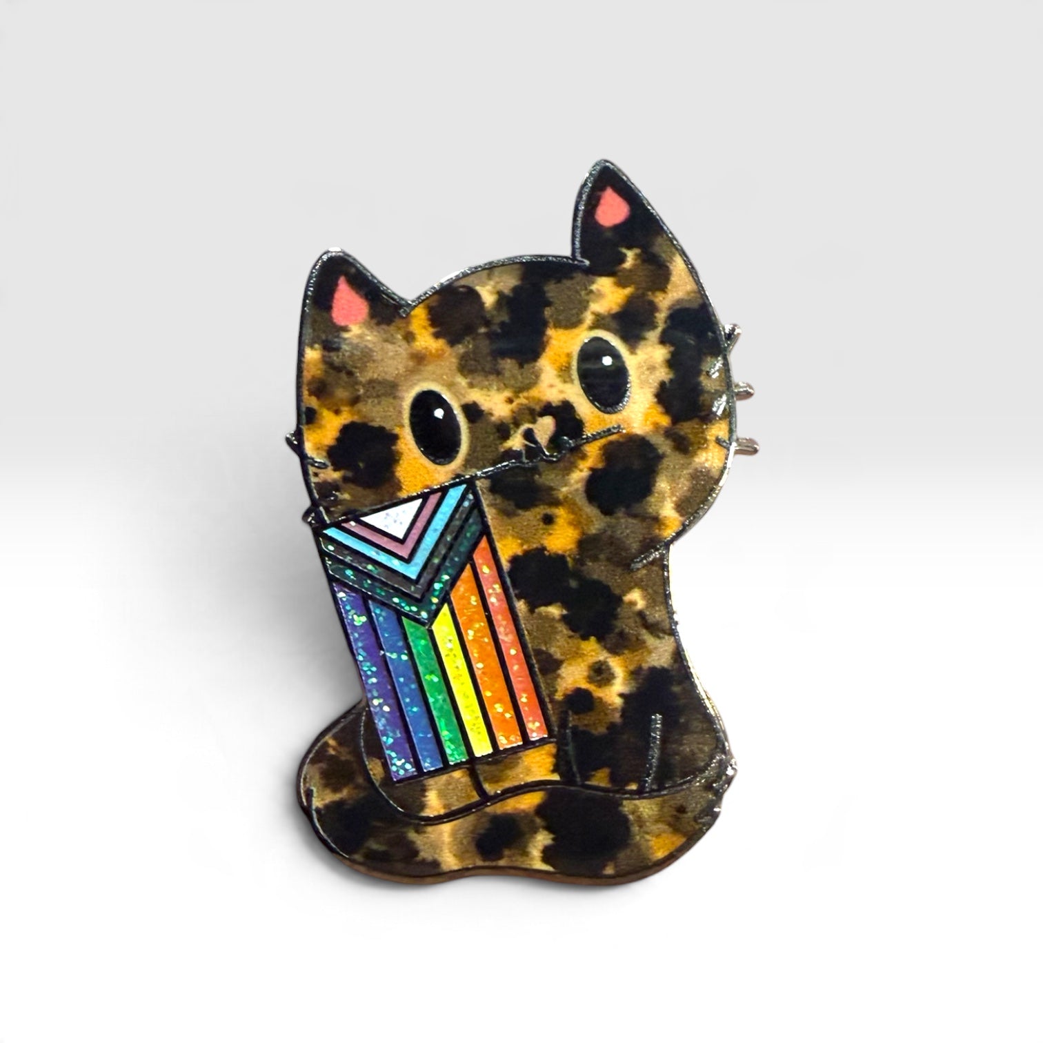 A small cat-shaped pin with a black and tan leopard print, holding a colorful striped flag  with rainbow stripes, photographed on a clean light gray background.