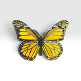 A butterfly-shaped pin with vibrant yellow wings outlined in black and detailed with metallic gold-toned patterns, set against a clean light gray background.