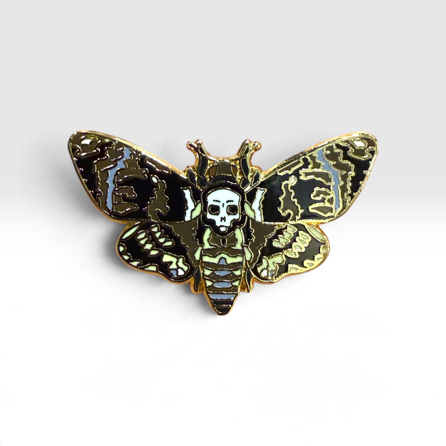 A gothic moth enamel pin featuring a skull and crossbones motif, with black, gold, and light blue patterned wings on a white background.