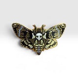 A gothic moth enamel pin featuring a skull and crossbones motif, with black, gold, and light blue patterned wings on a white background.