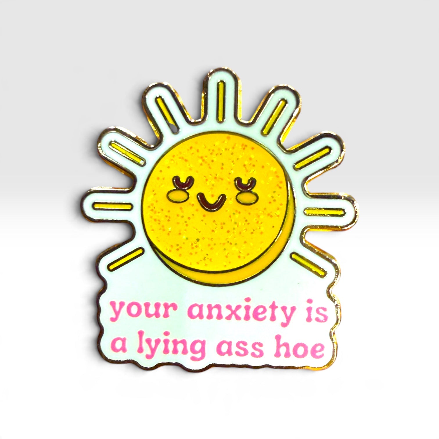 A die-cut enamel pin featuring a yellow glittery sun with a smiling face and the text 'your anxiety is a lying ass hoe' in pink lowercase letters, shown on a light gray gradient background.