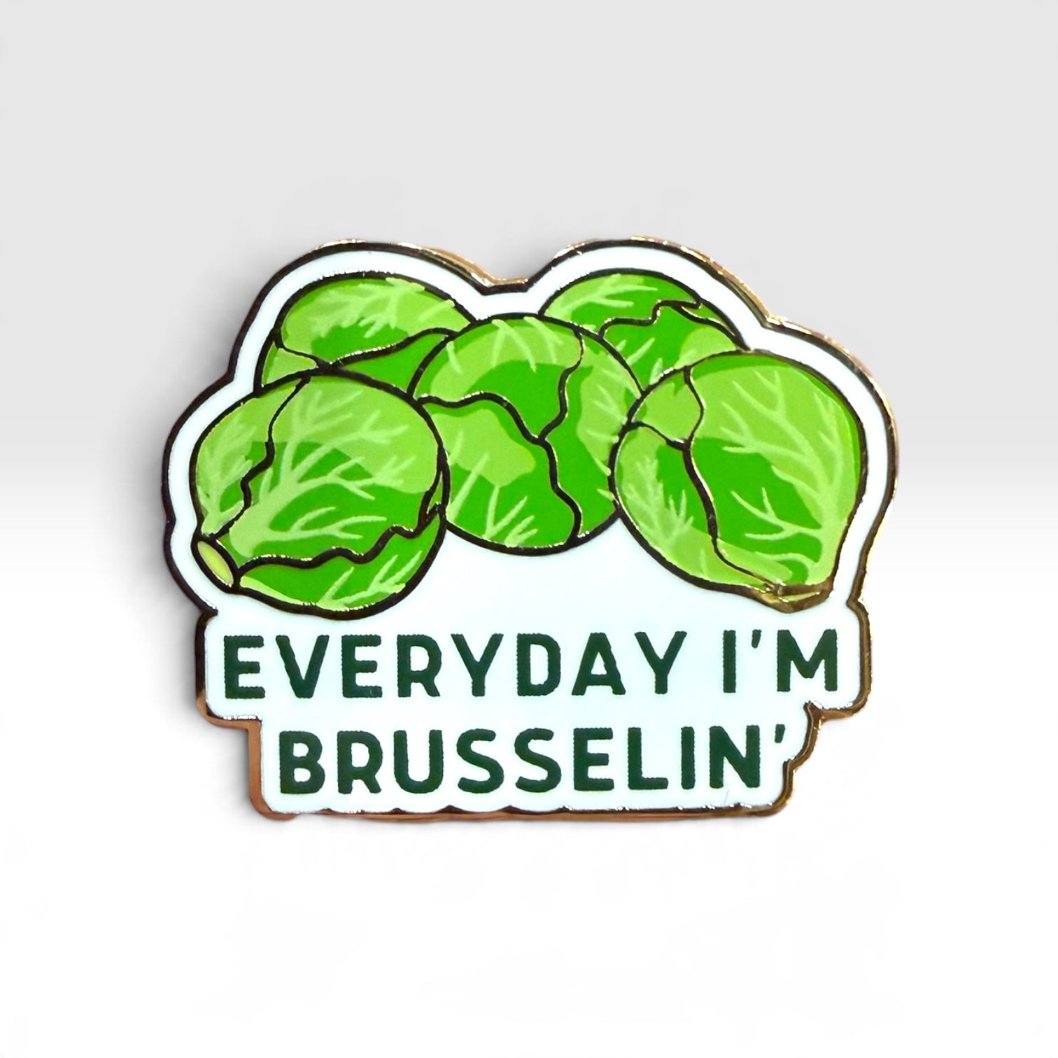 A die-cut enamel pin featuring green Brussels sprouts on a white background with the text 'EVERYDAY I'M BRUSSELIN'' in dark green lettering, outlined in gold-toned metal, shown on a light gray gradient background.