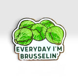 A die-cut enamel pin featuring green Brussels sprouts on a white background with the text 'EVERYDAY I'M BRUSSELIN'' in dark green lettering, outlined in gold-toned metal, shown on a light gray gradient background.
