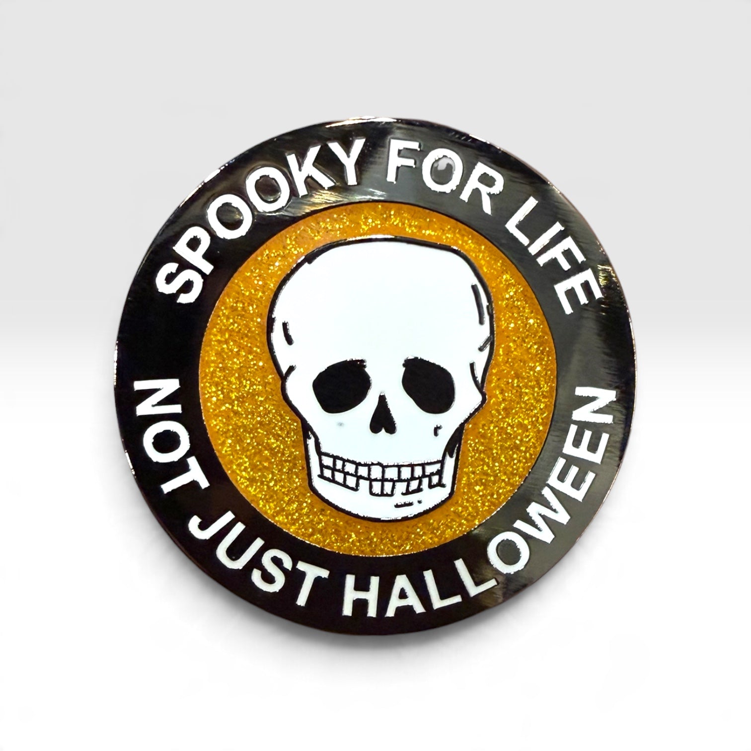 A circular Halloween-themed enamel pin with a black outer ring, white skull in the center, and glittery gold background. The text reads 'SPOOKY FOR LIFE' and 'NOT JUST HALLOWEEN'.