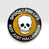 A circular Halloween-themed enamel pin with a black outer ring, white skull in the center, and glittery gold background. The text reads 'SPOOKY FOR LIFE' and 'NOT JUST HALLOWEEN'.