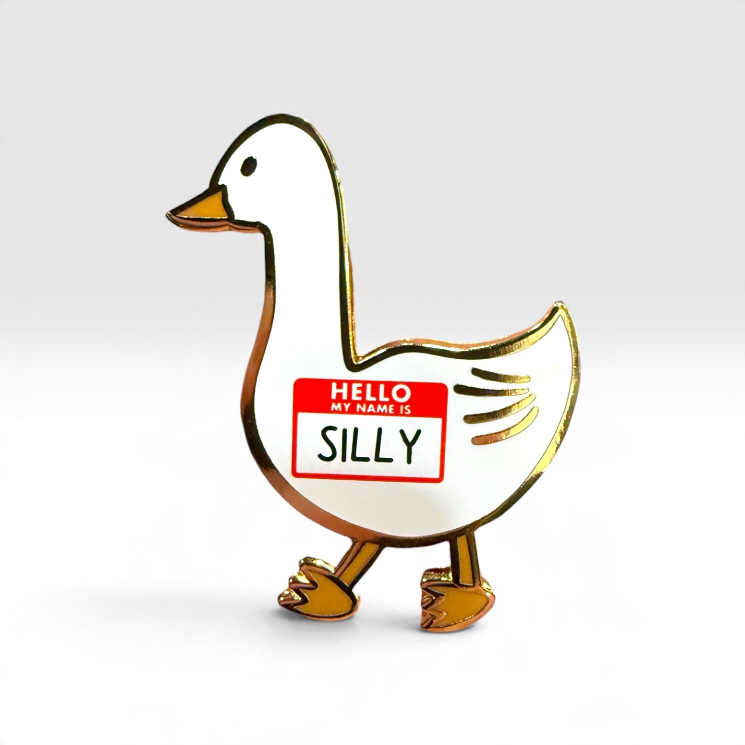 A white goose-shaped enamel pin with gold metal outlines, featuring a red and white 'HELLO MY NAME IS SILLY' name tag on its side, set against a clean light gray background.