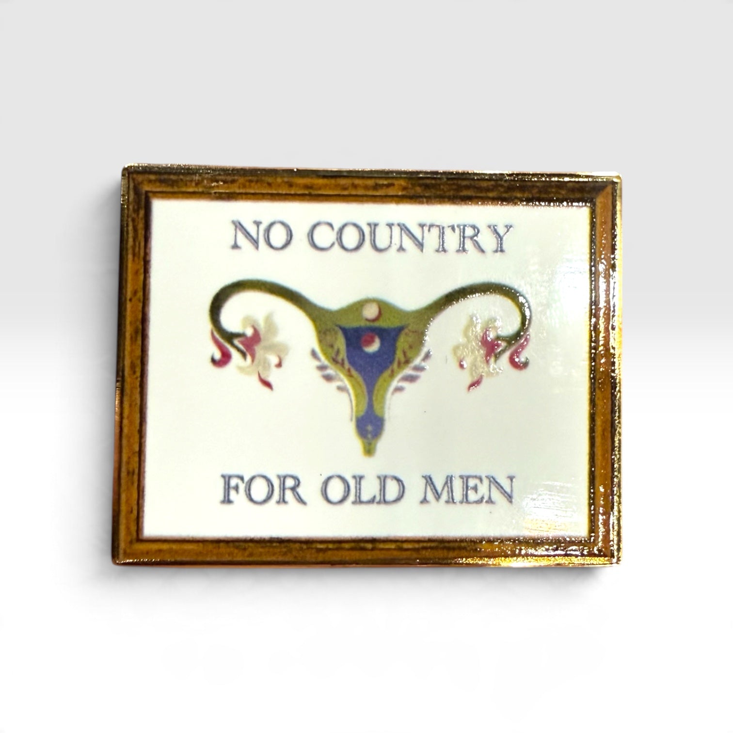 A rectangular enamel pin with a gold-toned frame and a white face, featuring the text 'NO COUNTRY FOR OLD MEN' in blue-gray lettering around a stylized uterus illustration in blue, gold, and pink.