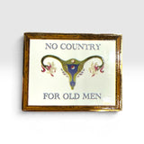A rectangular enamel pin with a gold-toned frame and a white face, featuring the text 'NO COUNTRY FOR OLD MEN' in blue-gray lettering around a stylized uterus illustration in blue, gold, and pink.