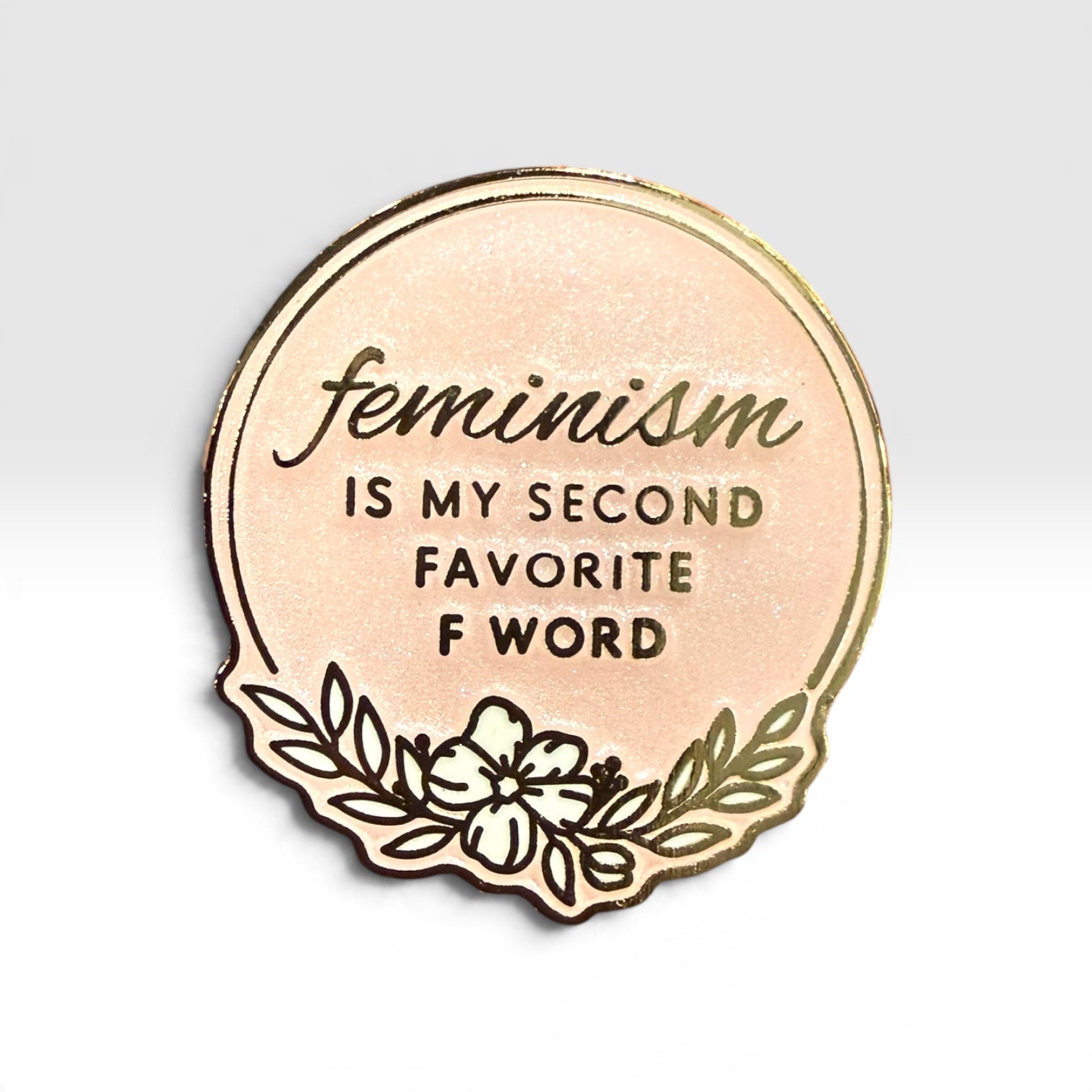 A circular enamel pin with a rose gold metallic background and gold-toned border, featuring the text 'feminism IS MY SECOND FAVORITE F WORD' in dark lettering, with a floral line-art design at the bottom, shown on a light gray gradient background.