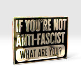 A rectangular enamel pin with a black background and metallic gold-toned lettering that reads, 'IF YOU'RE NOT ANTI-FASCIST WHAT ARE YOU?' on a light gray studio background.
