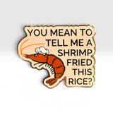 A novelty enamel pin shaped like a shrimp with a chef's hat, featuring the text 'YOU MEAN TO TELL ME A SHRIMP FRIED THIS RICE?' in black lettering on a light beige background with a gold-toned outline.
