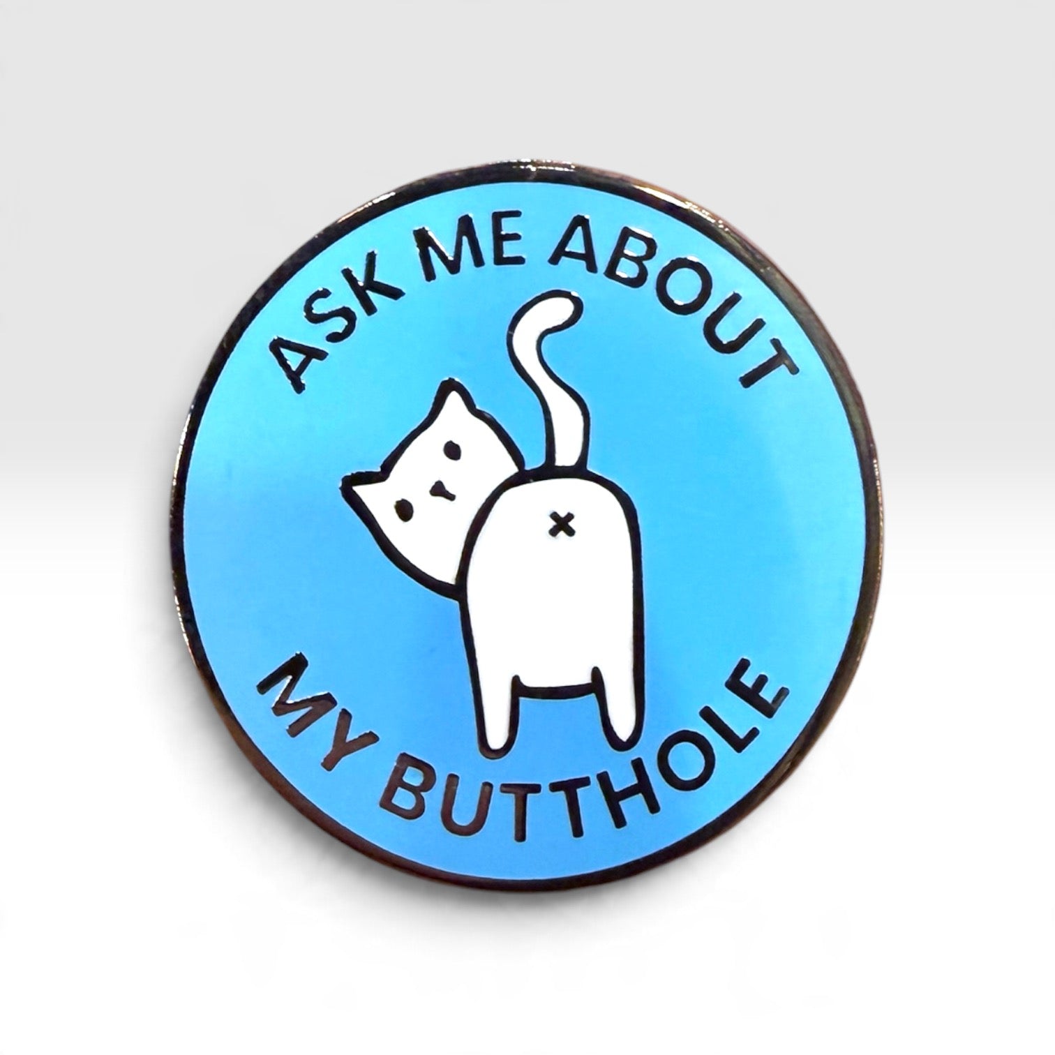 A round enamel pin with a light blue background, featuring a white cartoon cat with its back turned and the text 'ASK ME ABOUT MY BUTTHOLE' in black lettering.