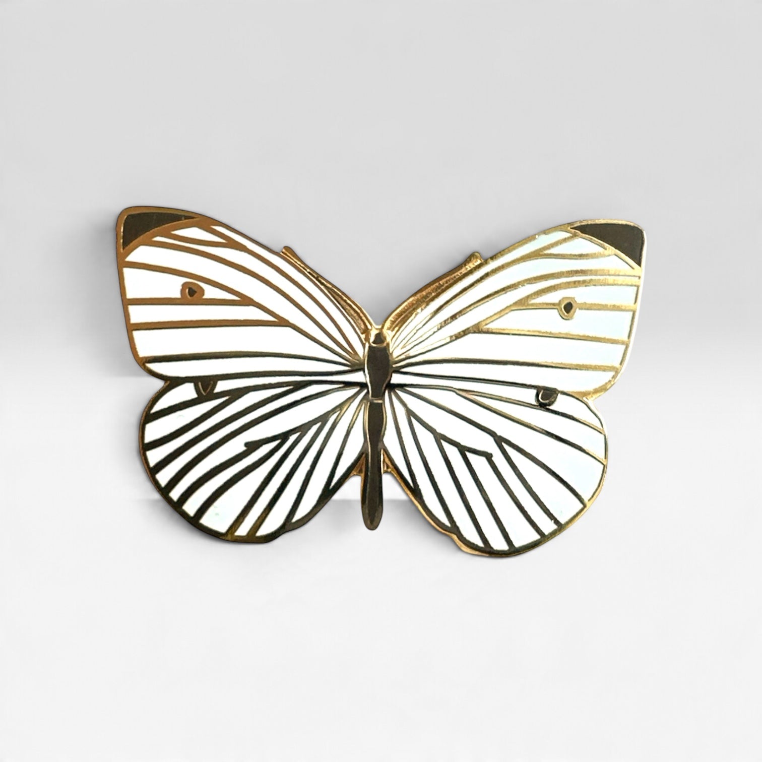 A butterfly-shaped pin with white enamel wings outlined in gold and black striped detailing, shown against a light gray background.