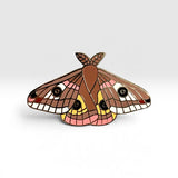 A detailed enamel pin shaped like a moth, featuring brown wings with pink, yellow, and white patterns and black eyes, set against a clean light gray background.