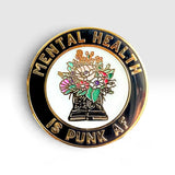 A circular enamel pin with a black outer ring, gold lettering that reads 'MENTAL HEALTH IS PUNK AF', and a central illustration of black boots holding a colorful bouquet of flowers on a white background.