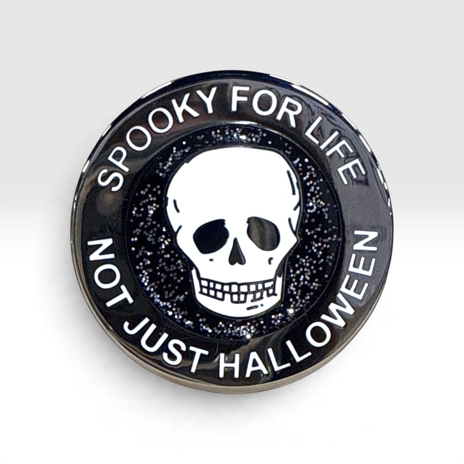 A round black enamel pin featuring a white skull in the center, surrounded by the text 'SPOOKY FOR LIFE' and 'NOT JUST HALLOWEEN' on a light gray background.