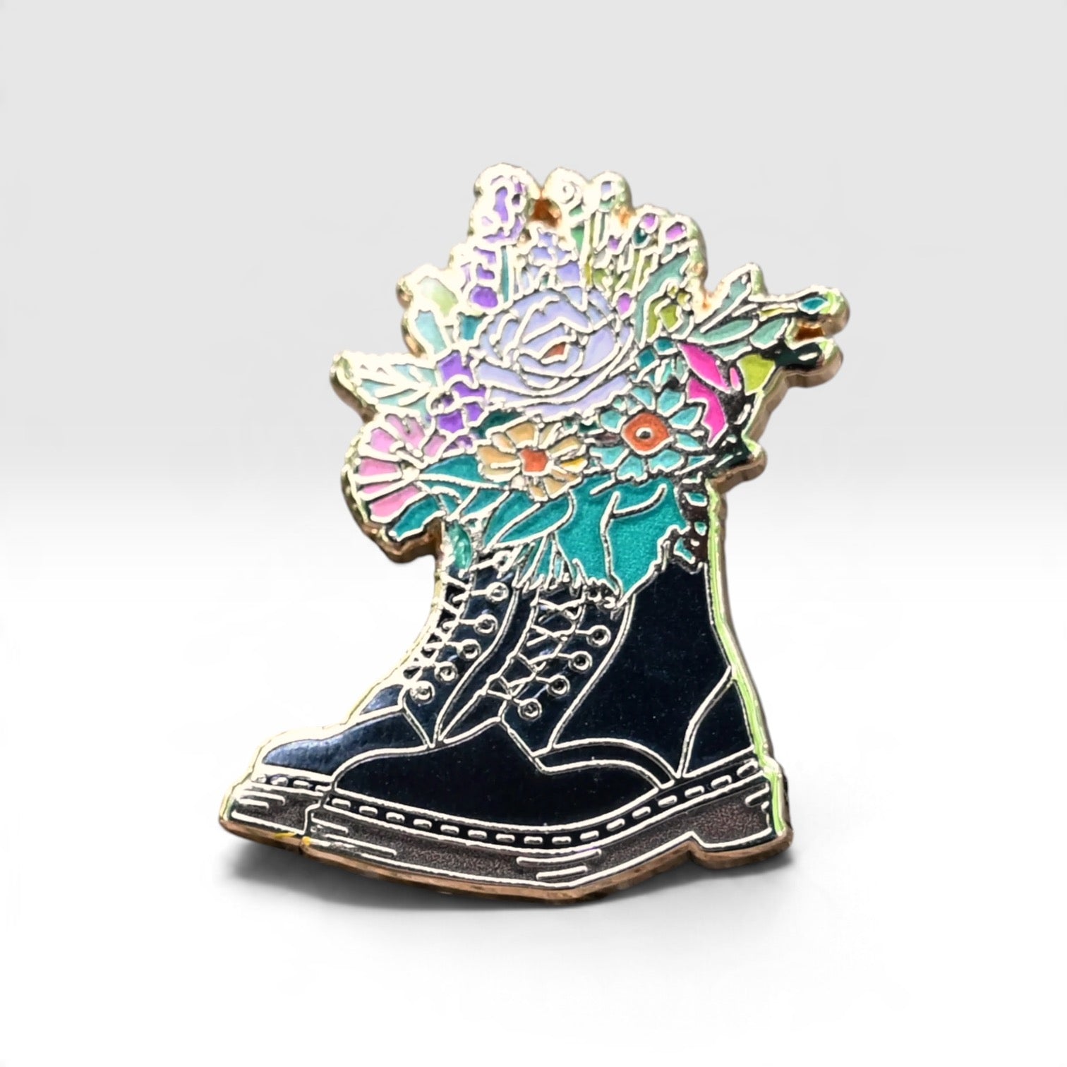 An enamel pin shaped like a black lace-up boot with a colorful floral bouquet growing from the top, set against a plain light gray background.