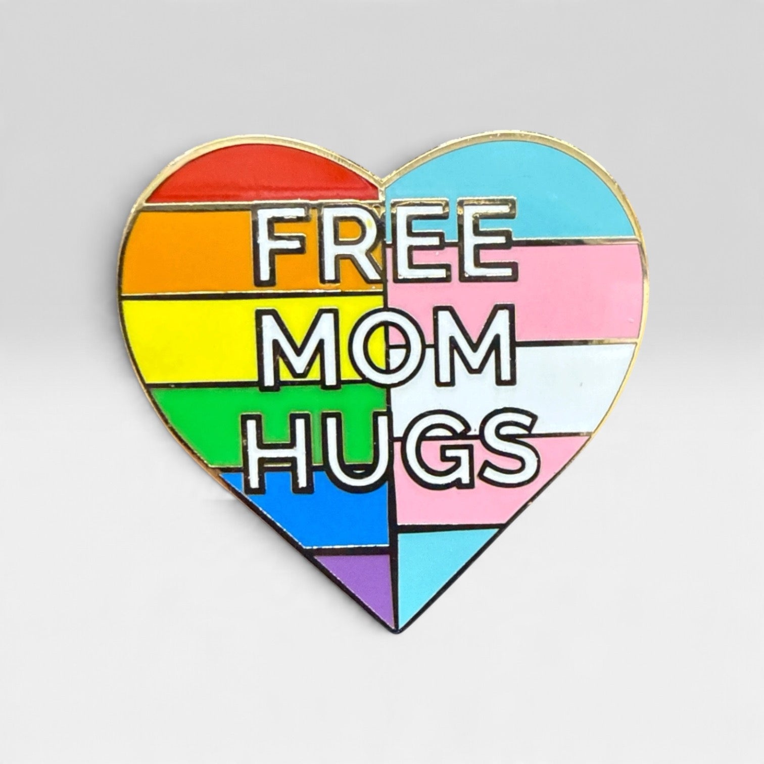 A heart-shaped enamel pin with a rainbow-striped design and the words 'FREE MOM HUGS' in bold white letters with black outlines, set against a light gray background.