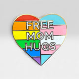 A heart-shaped enamel pin with a rainbow-striped design and the words 'FREE MOM HUGS' in bold white letters with black outlines, set against a light gray background.