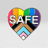 A heart-shaped enamel pin featuring the word 'SAFE' in bold white letters over a rainbow-striped background and a Progress Pride flag motif at the bottom, shown on a light gray background.