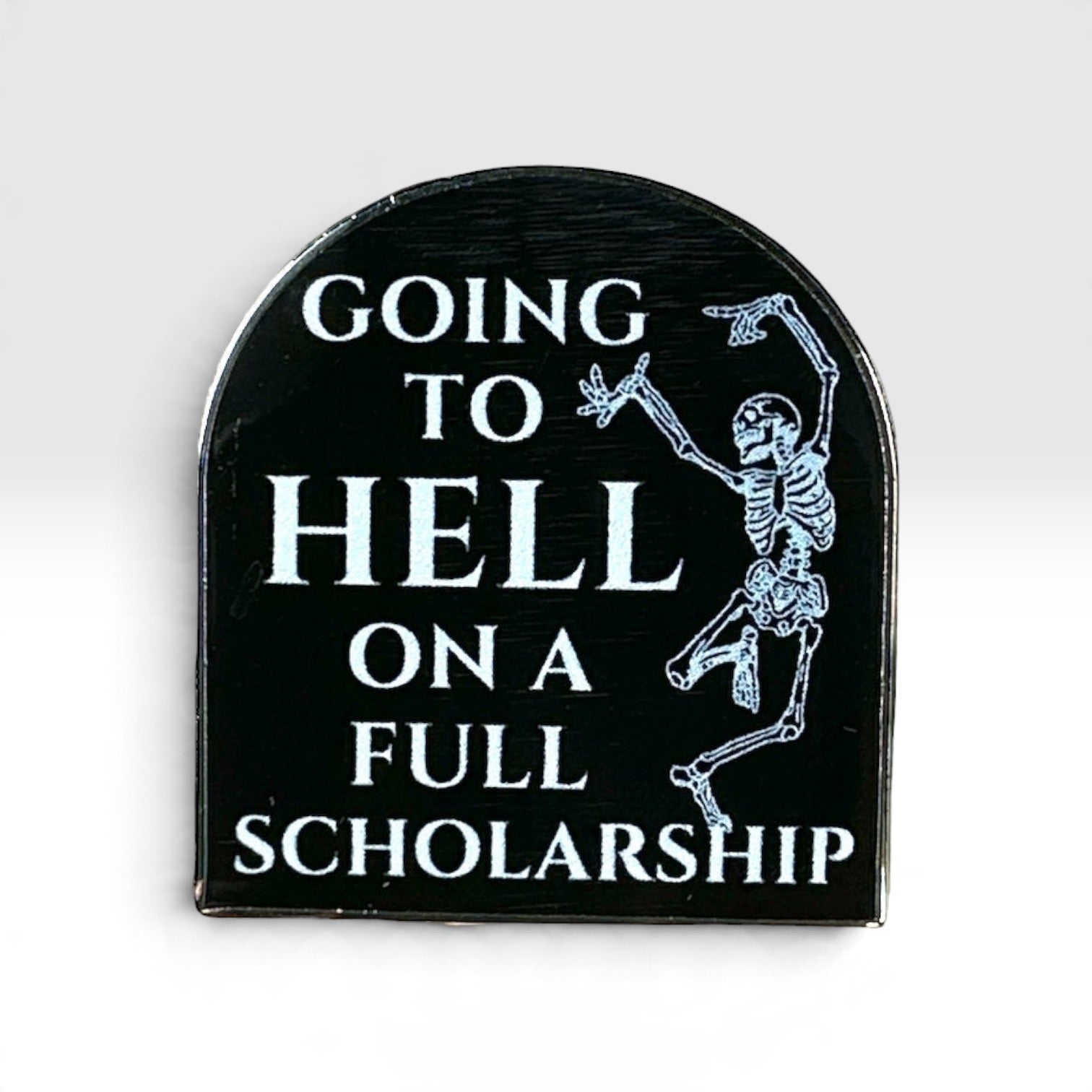 A tombstone-shaped black enamel pin featuring white text that reads 'GOING TO HELL ON A FULL SCHOLARSHIP' and a white skeleton dancing on the right side, shown against a light gray gradient background.