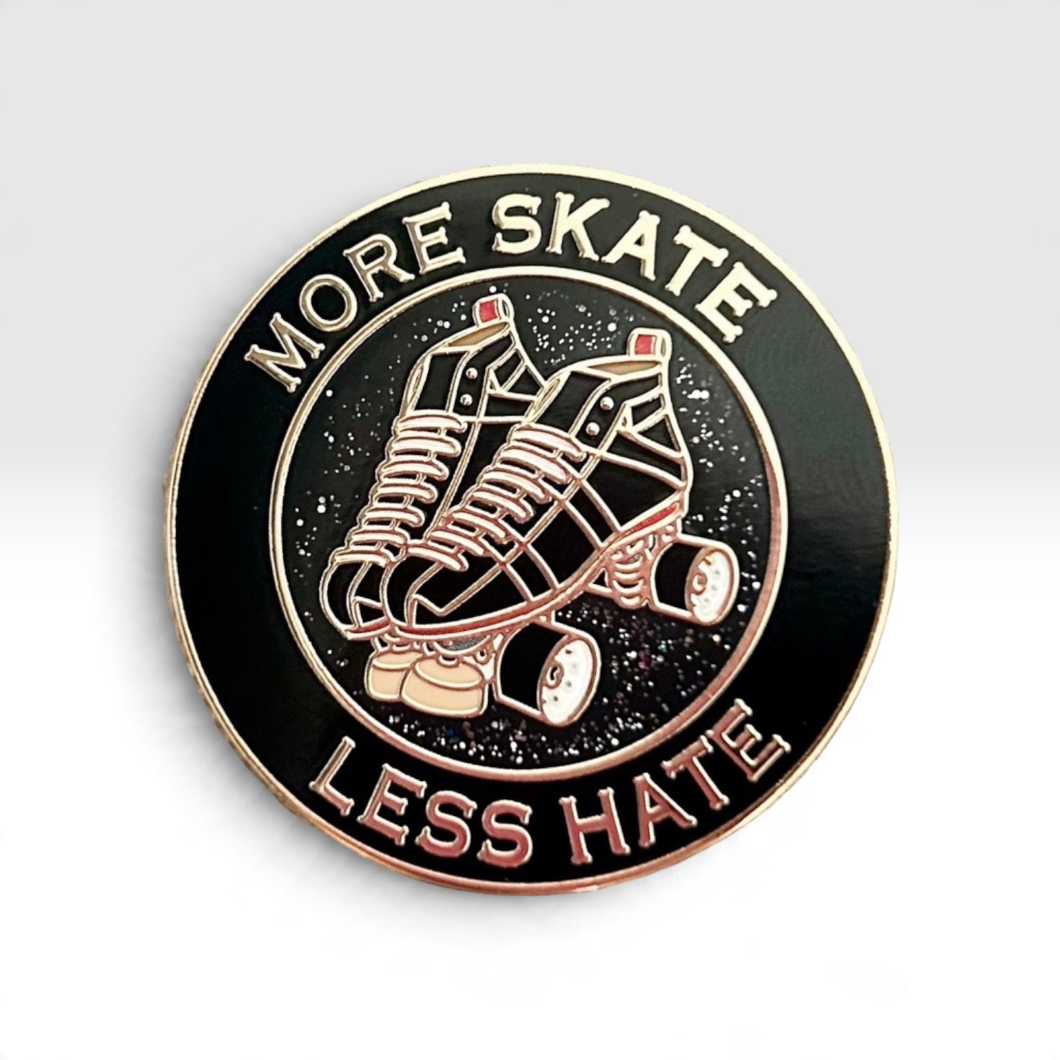 A circular enamel pin with roller skate graphics and the text 'MORE SKATE LESS HATE' on a light gray gradient background.