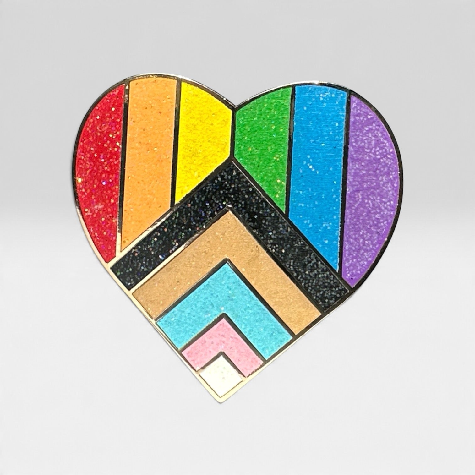 A heart-shaped enamel pin featuring the Progress Pride flag design with glittery stripes and chevron patterns in red, orange, yellow, green, blue, purple, black, brown, light blue, pink, and white, on a light gray background.