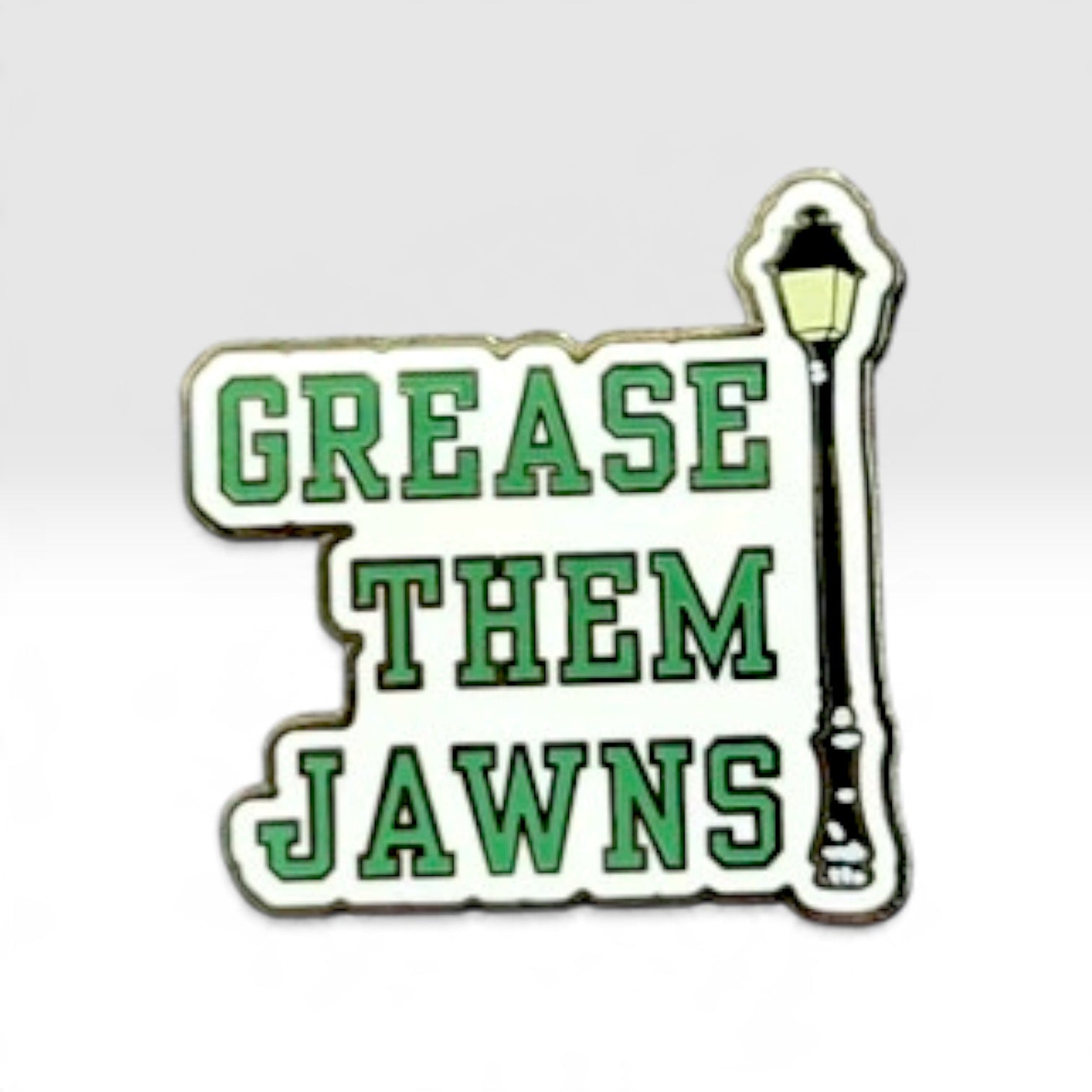 A novelty enamel pin featuring the phrase 'GREASE THEM JAWNS' in green block letters on a white background, with a black and yellow street lamp graphic on the right side.