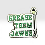 A novelty enamel pin featuring the phrase 'GREASE THEM JAWNS' in green block letters on a white background, with a black and yellow street lamp graphic on the right side.