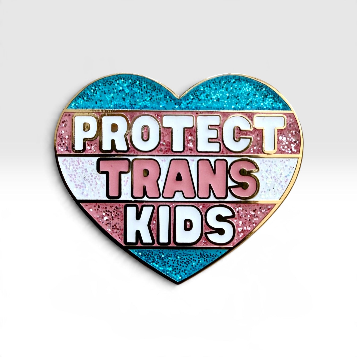 A heart-shaped enamel pin with the text 'PROTECT TRANS KIDS' in white and pink lettering over horizontal glitter stripes in blue, pink, white, and pink, outlined in gold.