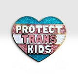 A heart-shaped enamel pin with the text 'PROTECT TRANS KIDS' in white and pink lettering over horizontal glitter stripes in blue, pink, white, and pink, outlined in gold.