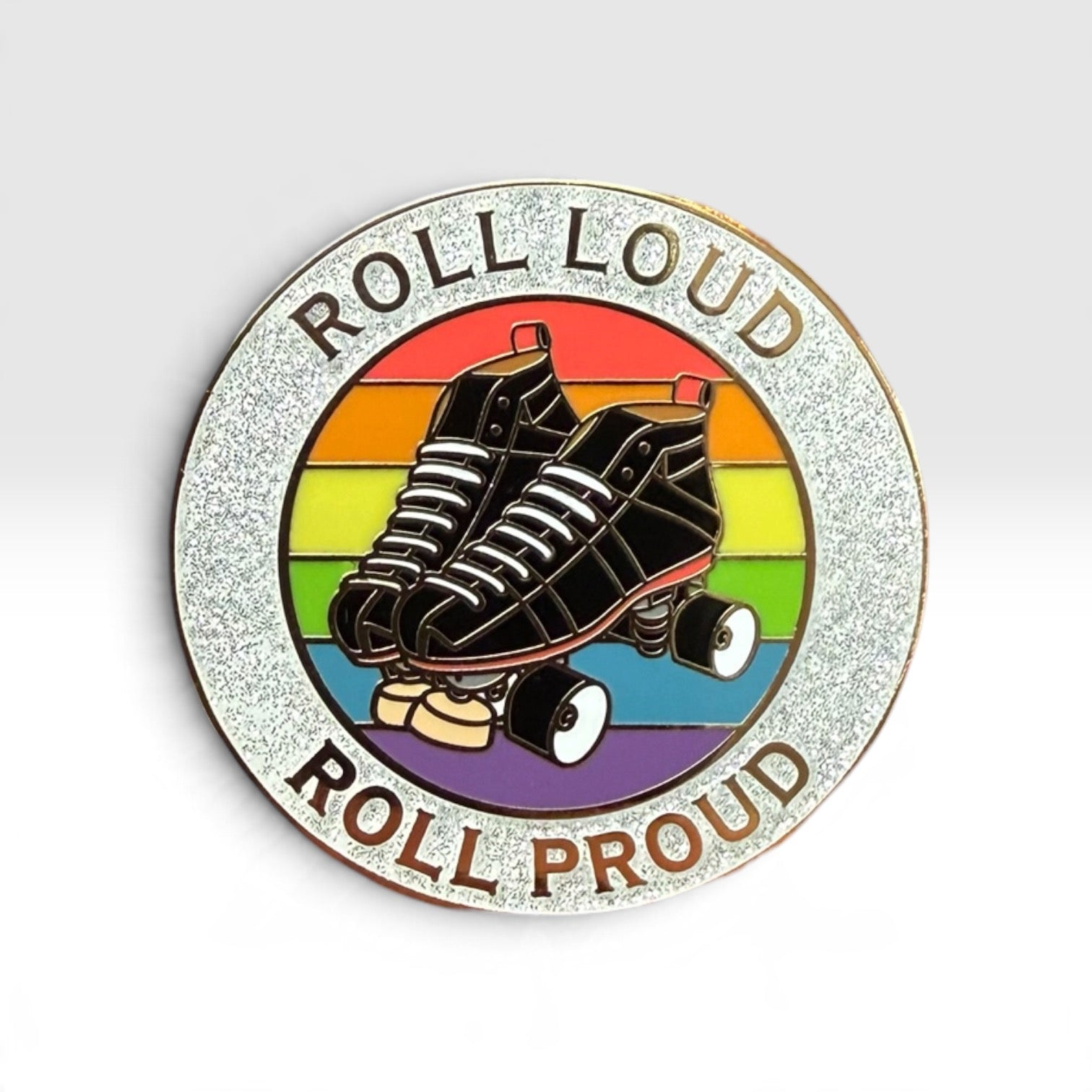 A circular enamel pin featuring roller skates over a retro rainbow background with the words 'ROLL LOUD' at the top and 'ROLL PROUD' at the bottom, shown on a light gray gradient background.