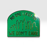 A green enamel pin with a gold-tone outline and text reading 'NO ONE LIKES US WE DON'T CARE' above and below a simple skyline silhouette, shown on a light gray background.
