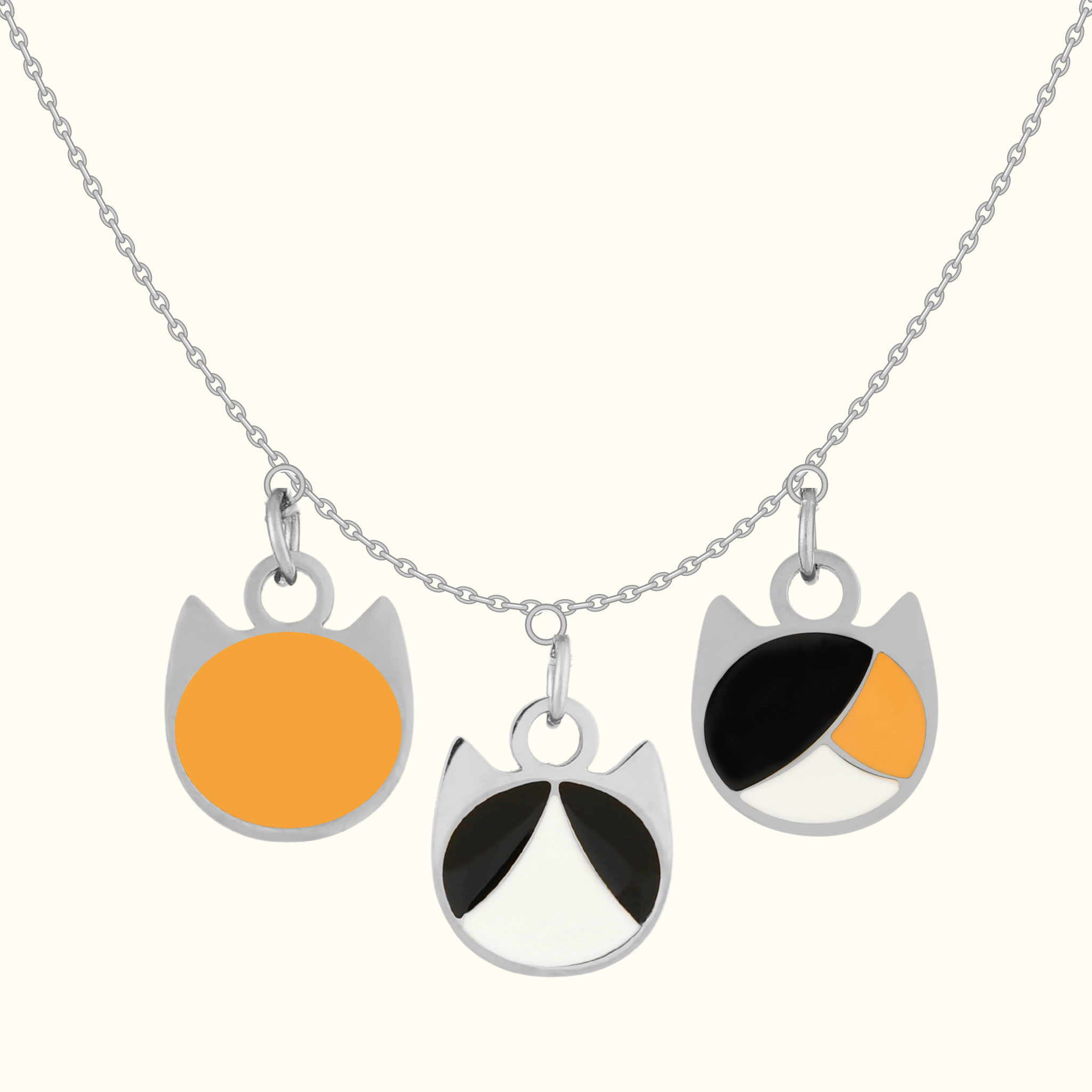 A silver chain necklace with three round cat-face style pendants in silver-tone metal with black, white, and orange enamel details, shown on a light cream background.