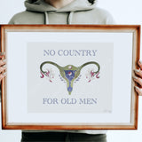A person in a light gray hoodie holds a framed art print featuring a stylized uterus illustration with the text 'NO COUNTRY FOR OLD MEN' in blue serif lettering. The print has a white mat and a warm brown wooden frame.